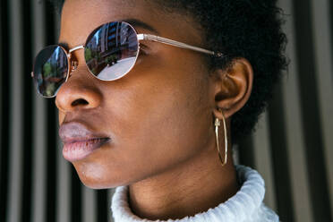 Seitenansicht einer jungen afroamerikanischen Frau mit modischem Pullover und Sonnenbrille, die im hellen Sonnenlicht vor schwarzem Hintergrund steht und wegschaut - ADSF27936