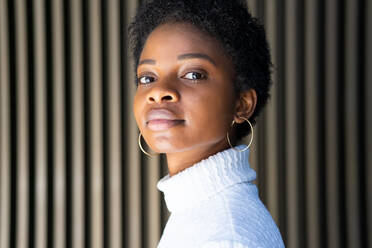 Side view of unemotional African American female in trendy sweater looking at camera against striped building wall on street - ADSF27938