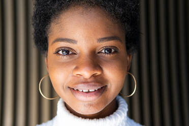 Happy stylish young black woman in crop sweater looking at camera while standing near striped building wall in city - ADSF27940