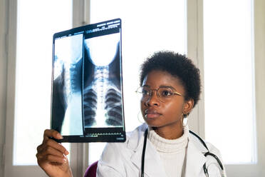 Junge schwarze Ärztin, die in der Nähe des Fensters steht und eine Röntgenaufnahme untersucht, während sie in einer Klinik arbeitet - ADSF27950