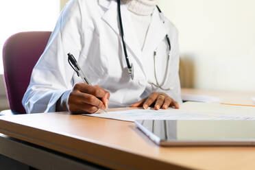 Nicht erkennbarer schwarzer Arzt mit Stethoskop, der Informationen auf ein Blatt Papier schreibt, während er einen medizinischen Bericht am Tisch im Büro einer modernen Klinik vorbereitet - ADSF27956