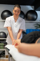 Happy masseuse in white robe massaging calf of crop female patient during physiotherapy session in clinic - ADSF27970