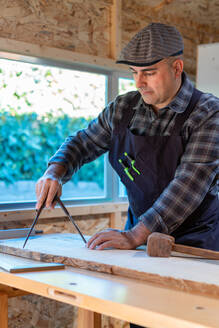 Männlicher Holzarbeiter, der einen professionellen Zirkel oder Teiler benutzt, während er ein Holzbrett an einer Werkbank in einer Tischlerwerkstatt anzeichnet - ADSF27983