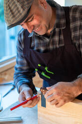 Positiver professioneller Holzarbeiter mit Bleistift und Messwerkzeug, der Markierungen auf einer Holzplatte anbringt, während er ein handwerkliches Objekt in einer Tischlerwerkstatt herstellt - ADSF27984