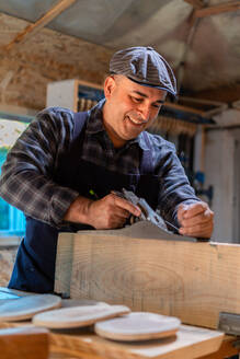 Gut gelaunter männlicher Tischler, der in einer professionellen Tischlerwerkstatt Holzteile mit einem Hobel glättet - ADSF27987