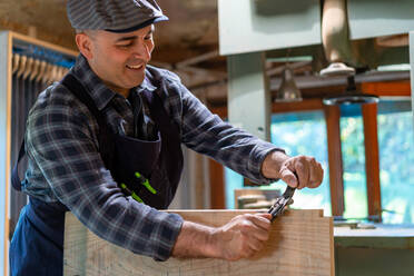 Fröhlicher männlicher Tischler beim Glätten von Holzbrettern mit einem professionellen Holzbearbeitungsmesser, einer manuellen Hobelmaschine in einer Werkstatt - ADSF27991