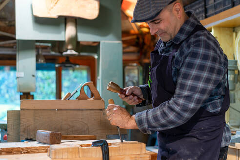 Seitenansicht eines Holzarbeiters mit Hammer und Meißel beim Schnitzen von Holzdetails in einer professionellen Tischlerwerkstatt - ADSF27992
