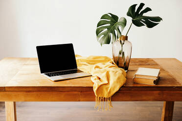 Moderner Arbeitsplatz mit Laptop und Büchern auf einem Holztisch mit grünem Laub in einer Glasvase und gelbem Tuch an einer weißen Wand - ADSF27995