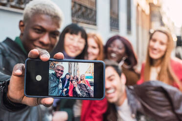 African American male taking selfie with smartphone with company of multiracial friends standing on street together - ADSF28023