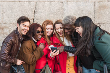 Company of multiethnic stylish friends gathering in street and watching funny video on mobile phone together - ADSF28028