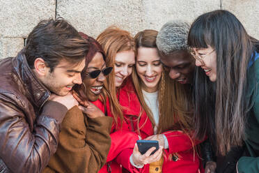 Company of multiethnic stylish friends gathering in street and watching funny video on mobile phone together - ADSF28029