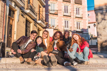 Company of content multiracial friends sitting in street and looking at camera on sunny day - ADSF28037