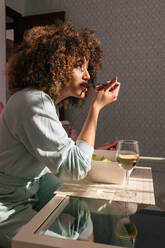 Side view of African American female eating salad while sitting at table with glass of wine and having tasty lunch at home - ADSF28066