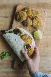 Draufsicht auf eine anonyme Person, die frische Süßkartoffel-Falafel und Salat in Tortilla über einem Schneidebrett auf einem Holztisch einwickelt - ADSF28085