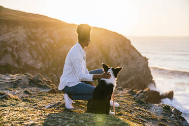 Seitenansicht einer jungen afroamerikanischen Besitzerin mit einem Border Collie Hund, die gemeinsam am Strand in der Nähe des wogenden Meeres bei Sonnenuntergang Zeit verbringen und die Aussicht betrachten - ADSF28094