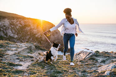 Junge afroamerikanische Besitzerin, die mit ihrem Border Collie Hund läuft, während sie gemeinsam Zeit am Strand in der Nähe des wogenden Meeres bei Sonnenuntergang verbringen - ADSF28096