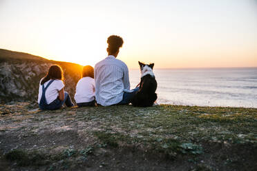 Back view of unrecognizable mother with children and Border Collie dog sitting on shore near sea and admiring sundown while spending evening together on beach - ADSF28097