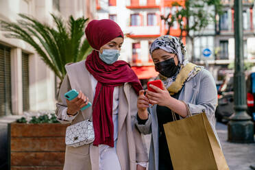 Muslimische Freundinnen in Hidschabs und mit Papiertüten, die nach dem Einkaufen auf der Straße ihr Smartphone benutzen - ADSF28098