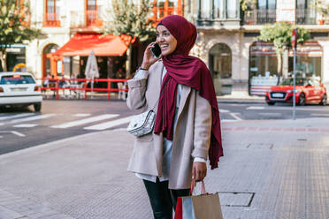 Fröhliche Muslimin im Hidschab und mit Einkaufstaschen, die die Straße entlangläuft und mit ihrem Smartphone spricht - ADSF28101