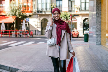 Fröhliche Muslimin im Hidschab und mit Einkaufstaschen, die die Straße entlangläuft und mit ihrem Smartphone spricht - ADSF28102