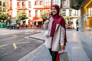 Fröhliche Muslimin im Hidschab und mit Einkaufstaschen, die die Straße entlangläuft und mit ihrem Smartphone spricht - ADSF28105