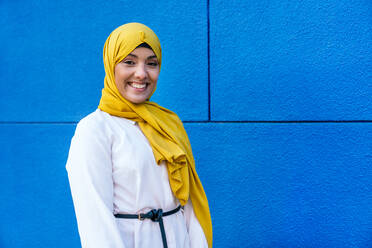 Seitenansicht einer trendigen muslimischen Frau in gelbem Hidschab, die auf der Straße auf blauem Hintergrund in die Kamera schaut - ADSF28121