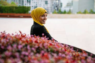 Side view of cheerful Muslim female in headscarf sitting on bench near flowerbed in street and looking at camera - ADSF28123