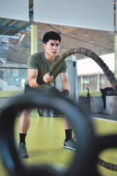 Konzentrierter asiatischer Mann beim Training mit dem Battle Rope in einem modernen Crossfit-Fitnessstudio - ADSF28124
