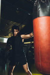 Junger fokussierter asiatischer Mann, der Boxen trainiert und Schläge ausführt, während er mit einem schweren Boxsack in einem modernen Fitnessstudio trainiert - ADSF28125