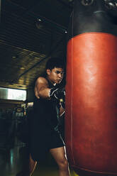 Junger fokussierter asiatischer Mann, der Boxen trainiert und Schläge ausführt, während er mit einem schweren Boxsack in einem modernen Fitnessstudio trainiert - ADSF28126