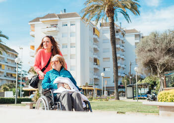 Adult woman pushing wheelchair with senior mother while walking on the street in city during summer - ADSF28130