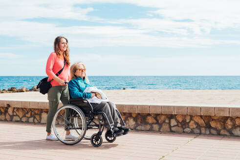 Seitenansicht einer erwachsenen Tochter, die mit ihrer älteren Mutter im Rollstuhl an der Strandpromenade entlang geht - ADSF28131