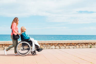 Side view of adult daughter walking with elderly mother in wheelchair along promenade near sea in summer - ADSF28132