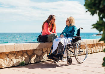 Adult woman and aged mother in wheelchair chilling on embankment and talking to each other on sunny day near sea - ADSF28135