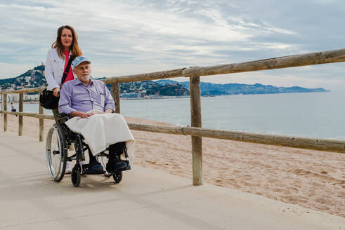 Erfreute erwachsene Tochter, die mit ihrem älteren Vater den Rollstuhl schiebt und den Spaziergang an der Promenade am Meer genießt - ADSF28137