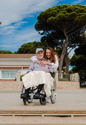 Lächelnde erwachsene Frau und älterer Mann im Rollstuhl entspannen zusammen in der Stadt an einem sonnigen Tag - ADSF28139