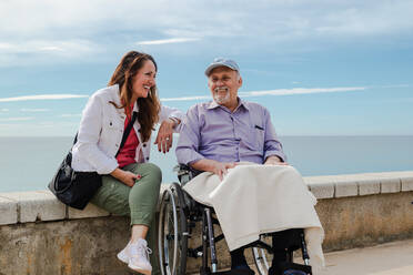 Zufriedene erwachsene Tochter und älterer Vater im Rollstuhl chillen im Sommer gemeinsam am Ufer des Meeres - ADSF28141