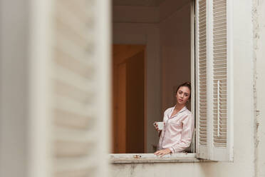 Tranquil female in pajama standing near window with cup of morning coffee and looking away - ADSF28171