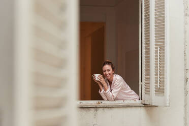 Tranquil female in pajama leaning in window with cup of morning coffee and looking at camera - ADSF28172