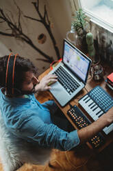 Von oben Seitenansicht eines fokussierten jungen Mannes mit Kopfhörern, der zu Hause an einem Tisch mit Synthesizer und Laptop arbeitet - ADSF28175