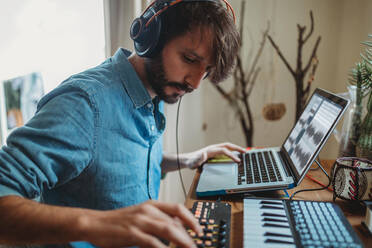 Seitenansicht eines jungen Mannes mit Kopfhörern, der einen Synthesizer und einen Laptop an einem Tisch zu Hause benutzt - ADSF28176