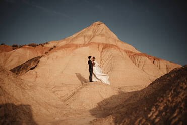 Side view of groom and bride embracing looking at each other near mountain against cloudy sundown sky in Bardenas Reales Natural Park in Navarra, Spain - ADSF28183