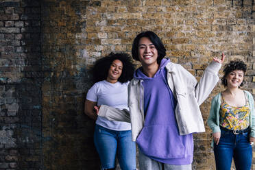 Smiling man gesturing while female friends standing in front of brick wall - ASGF00885