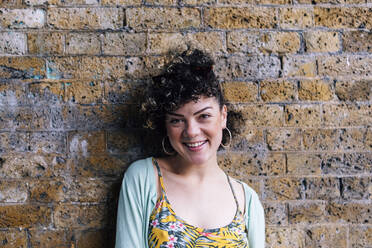 Smiling young woman with curly hair in front of brick wall - ASGF00892