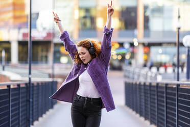 Cheerful woman with arms raised dancing while listening music through headphones on bridge - WPEF05067