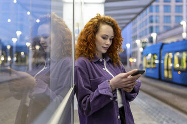 Young woman using mobile phone while leaning on glass at tram station - WPEF05074