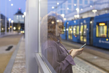 Frau mit Smartphone durch Glas an Straßenbahnhaltestelle gesehen - WPEF05076