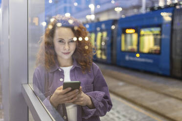 Schöne Frau mit Handy in der Hand, die durch Glas an einer Straßenbahnhaltestelle schaut - WPEF05077