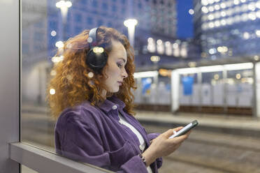 Frau mit Kopfhörern und Smartphone durch Glas an Straßenbahnhaltestelle gesehen - WPEF05080