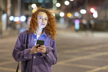 Rothaarige Frau mit Mobiltelefon, die nachts auf einer Kreuzung steht und wegschaut - WPEF05086